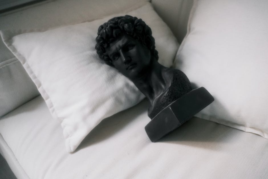 A black sculpture bust resting on white pillows, creating a contrast in a minimalist setting.
