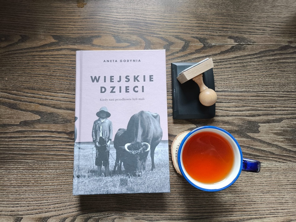 Książka Anety Godyni Wiejskie dzieci na tle surowego drewna, obok rzemieślnicza pieczątka ekslibris i kubek herbaty – recenzja na blogu Tradicon.