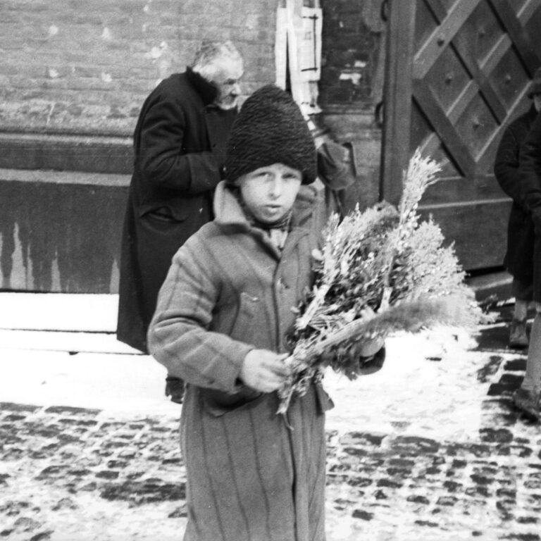 Czarno-białe zdjęcie z marca 1931 roku przedstawiające chłopca sprzedającego tradycyjne palmy wielkanocne na ulicy przed kościołem w Krakowie. W tle widać wiernych i architekturę kościoła.