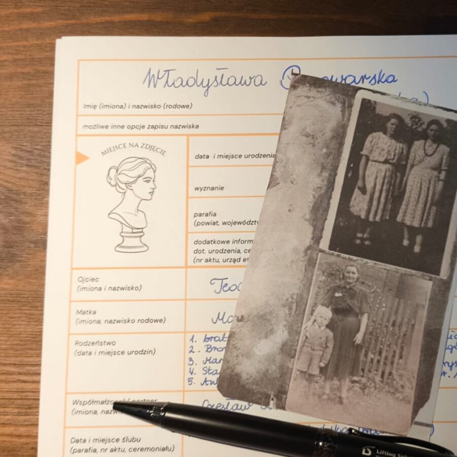Zdjęcie przedstawia formularz drzewa genealogicznego. Dokument leży na drewnianym blacie obok błękitnego notatnika ze starym, czarno-białym zdjęciem dwóch kobiet na okładce. Na pierwszym planie, na notatniku, spoczywa czarny długopis, gotowy do pisania. Kompozycja sugeruje proces dokumentowania historii rodziny i wspomnień.