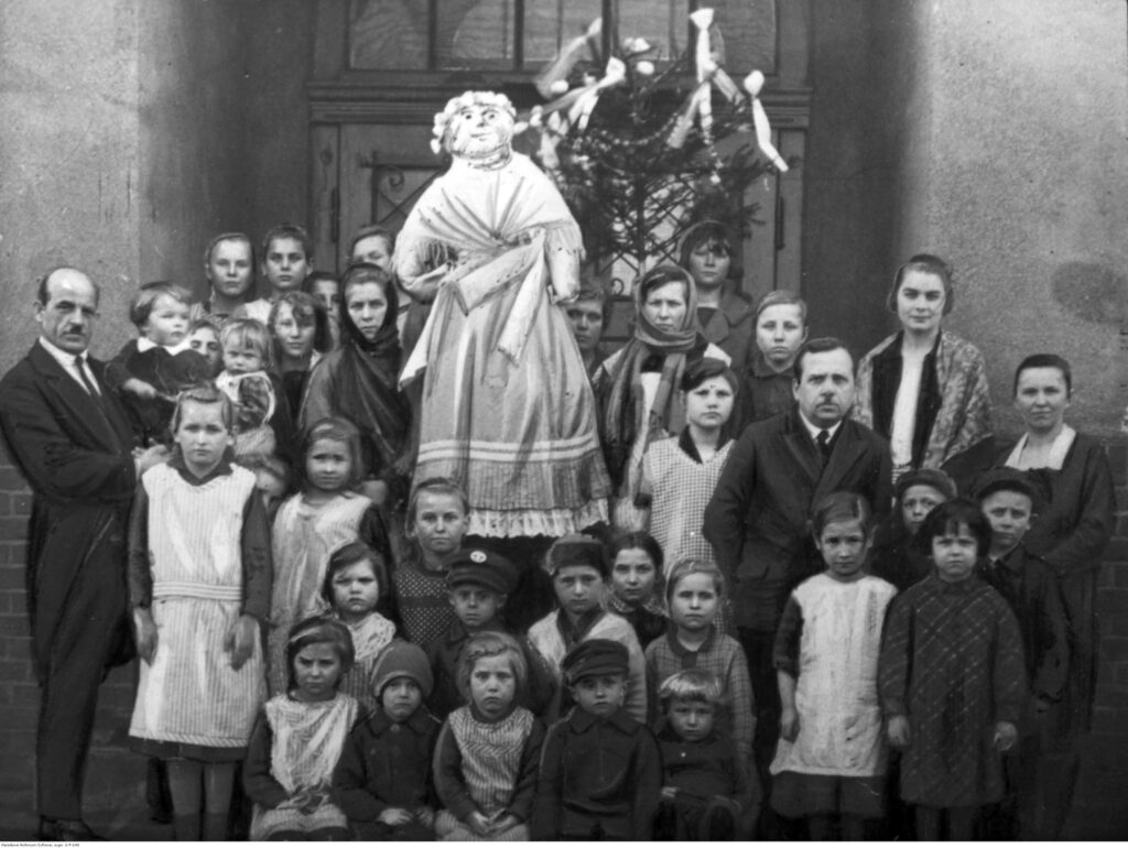 Czarno-białe zdjęcie archiwalne z lat 1925-1939 przedstawiające grupę dzieci w tradycyjnym pochodzie z kukłą Marzanny i przystrojonym Gaikiem Zielonym w miejscowości Repty.